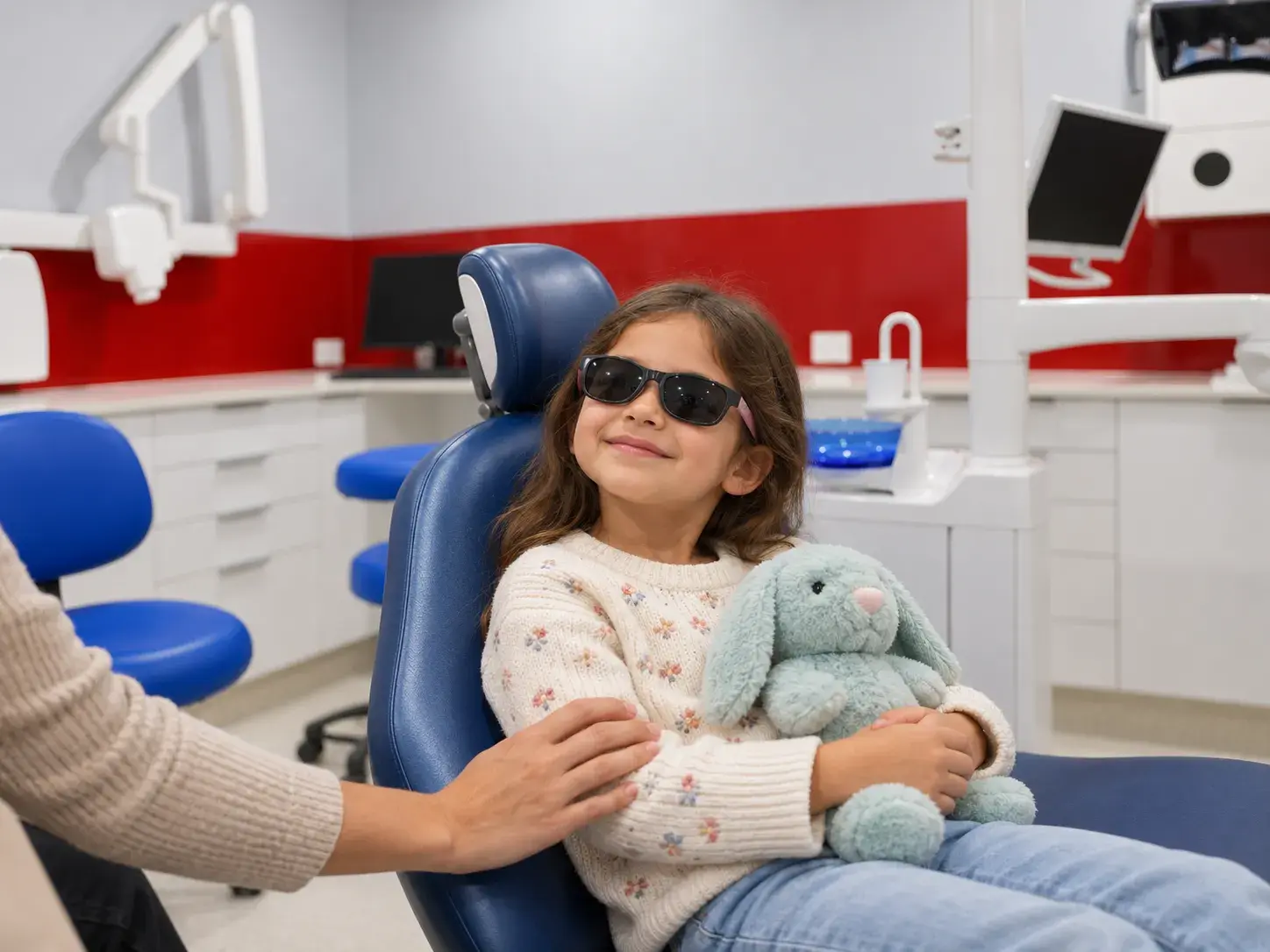 A young girl in the dental chair wearing sunglasses and holding a toy bunny
